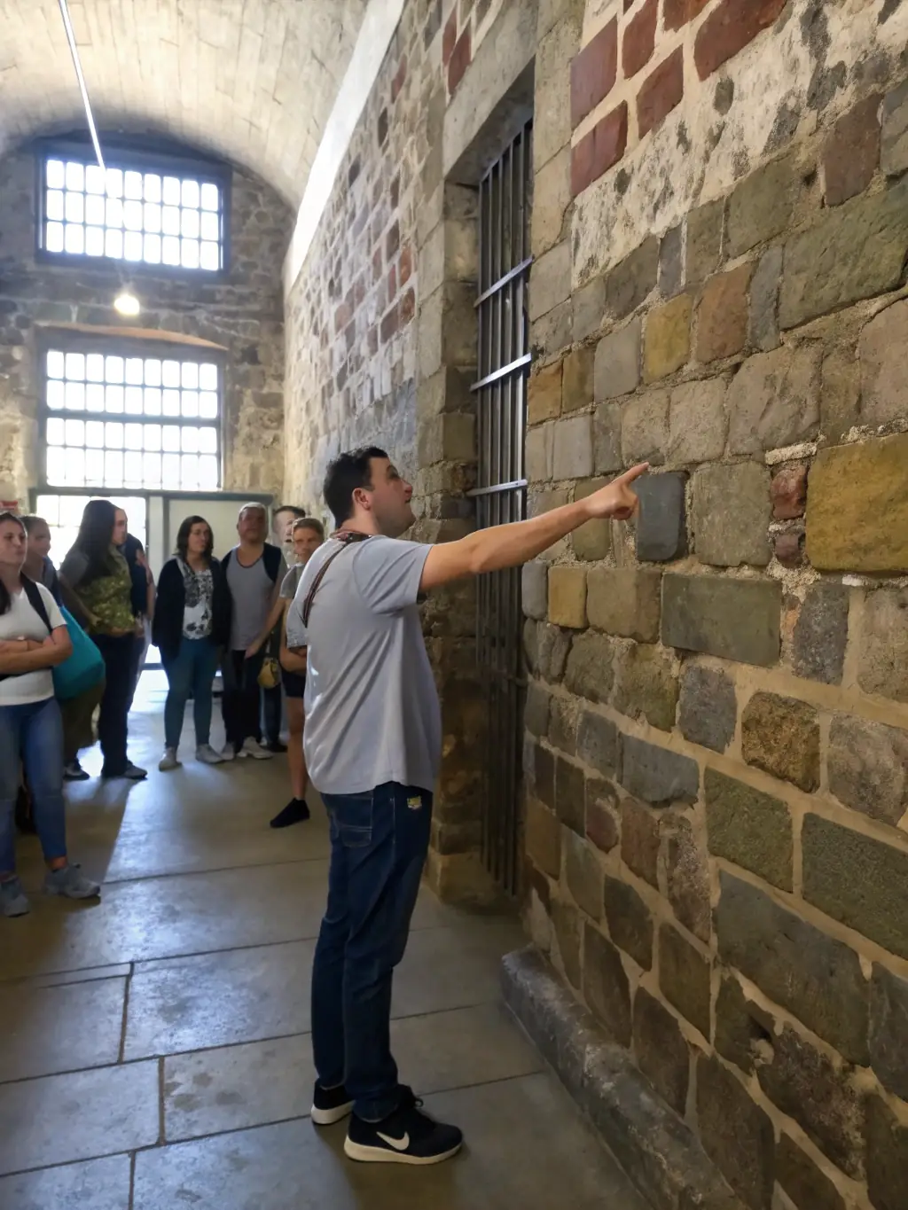 A close-up shot of a tour guide pointing out a historical detail on a cell wall, with visitors listening intently in the background.
